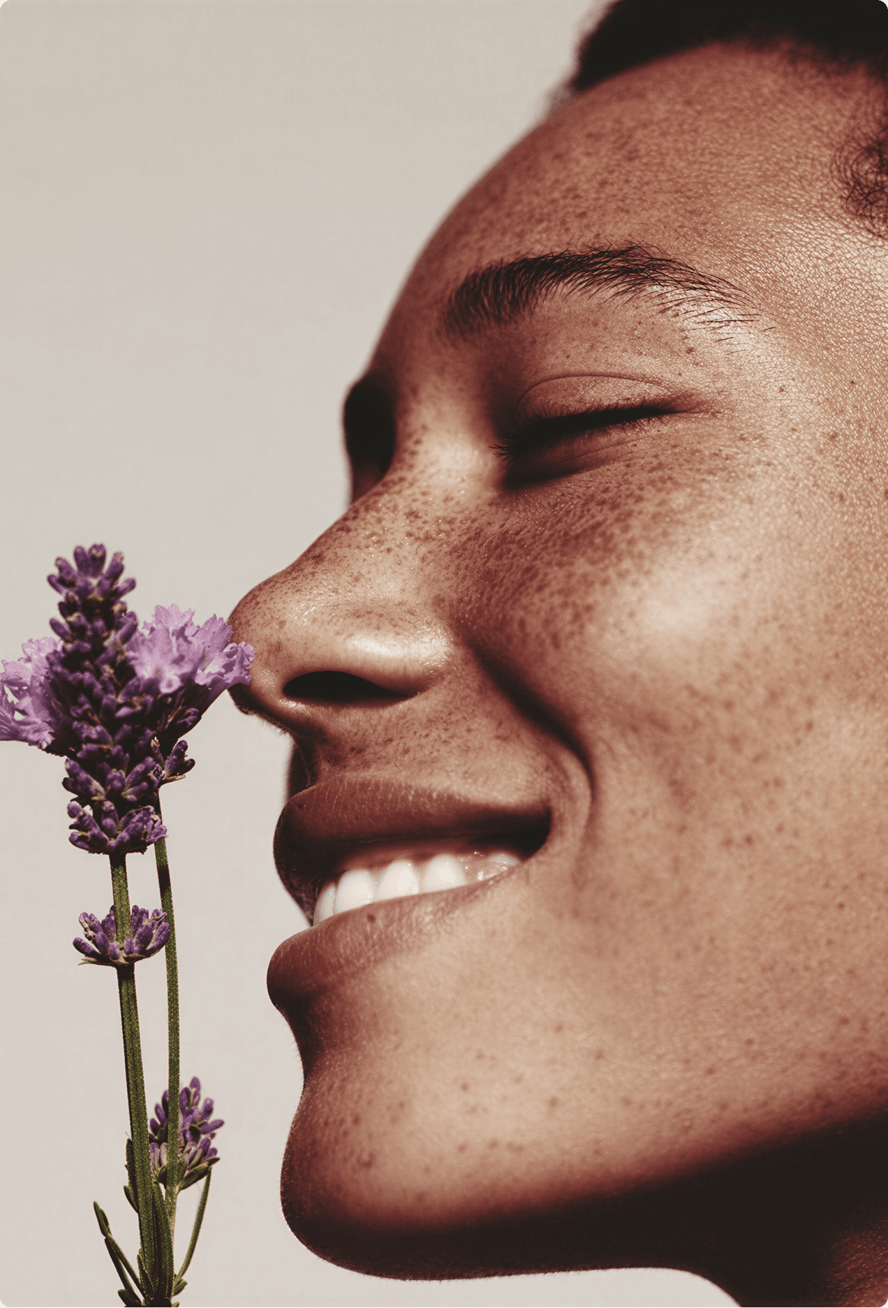 Close-up portrait of a smiling person inhaling the scent of fresh lavender stems against a neutral background