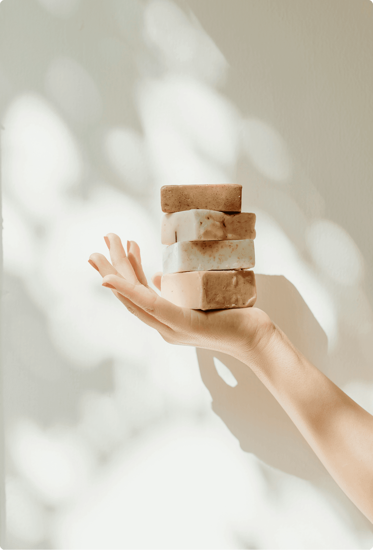 A hand balancing a stack of four handmade soap bars in natural tones against a soft white background with leaf shadows