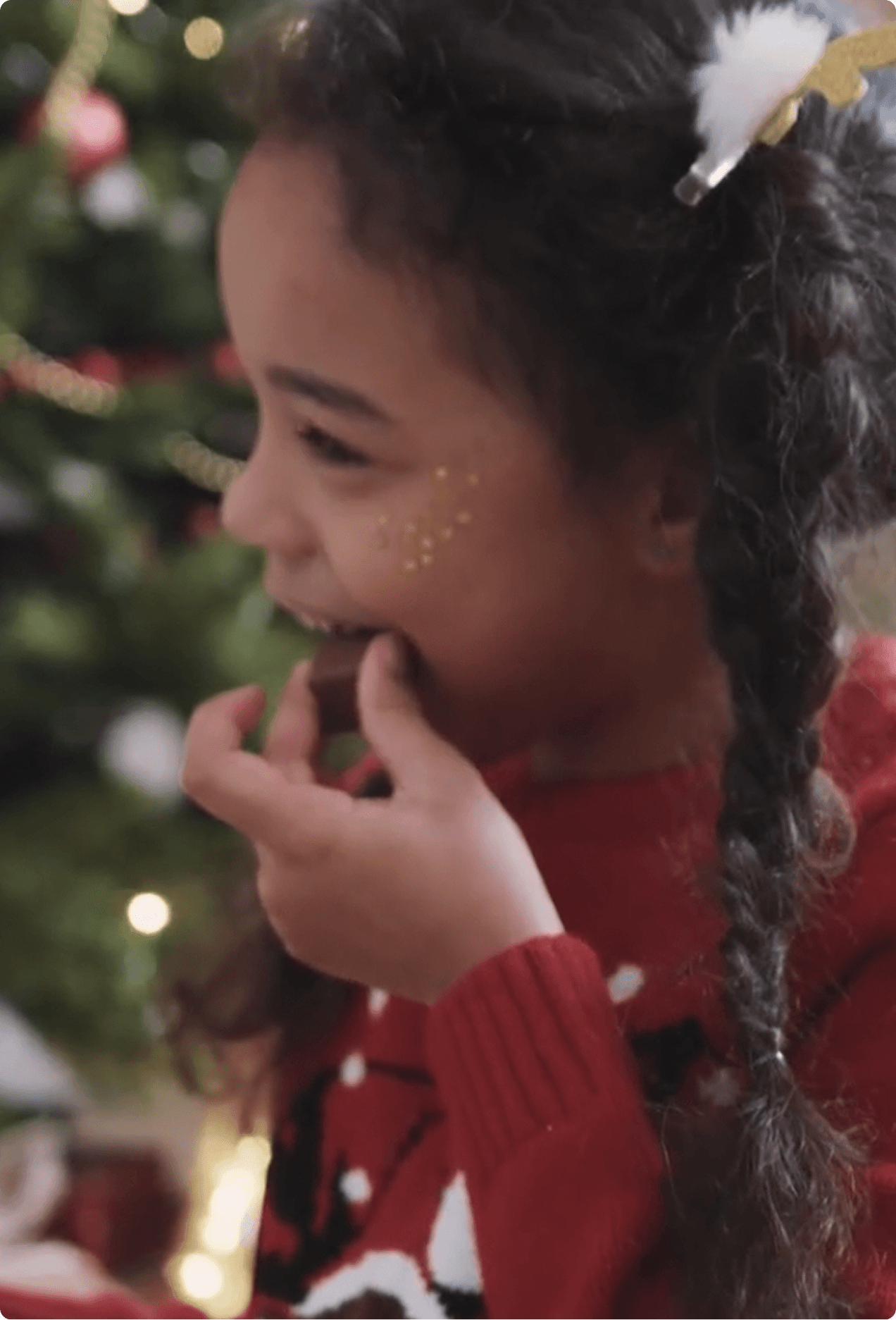Young girl in a red holiday sweater smiling while eating chocolate in front of a decorated Christmas tree