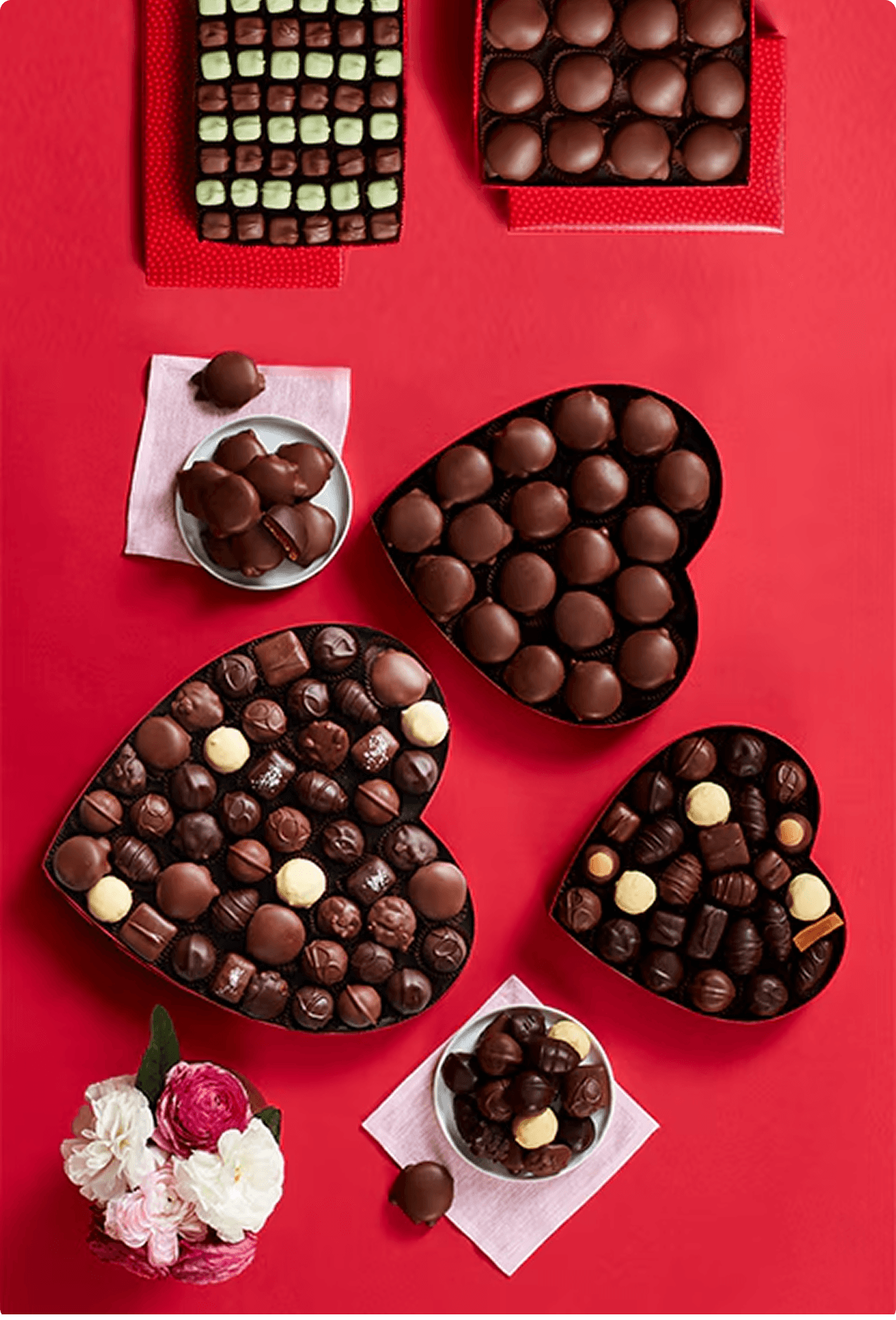 Overhead shot of Fannie May heart-shaped chocolate gift boxes in various sizes on a red surface with flowers and truffles