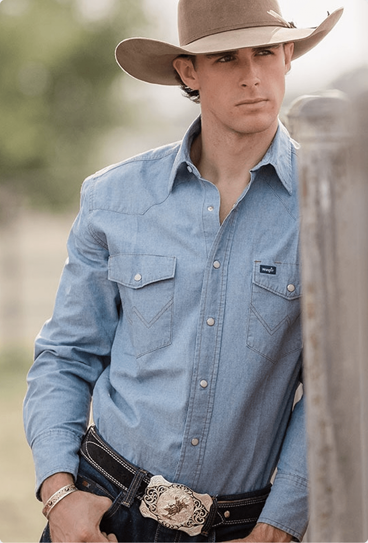 Young man in a cowboy hat and denim shirt wearing a Montana Silversmiths belt buckle, leaning against a weathered fence post