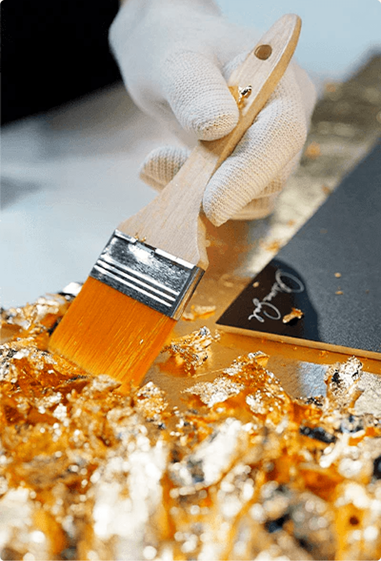 Gloved hand applying gold leaf flakes to an Oliver Gal canvas with a wide brush, with the brand signature visible on the work
