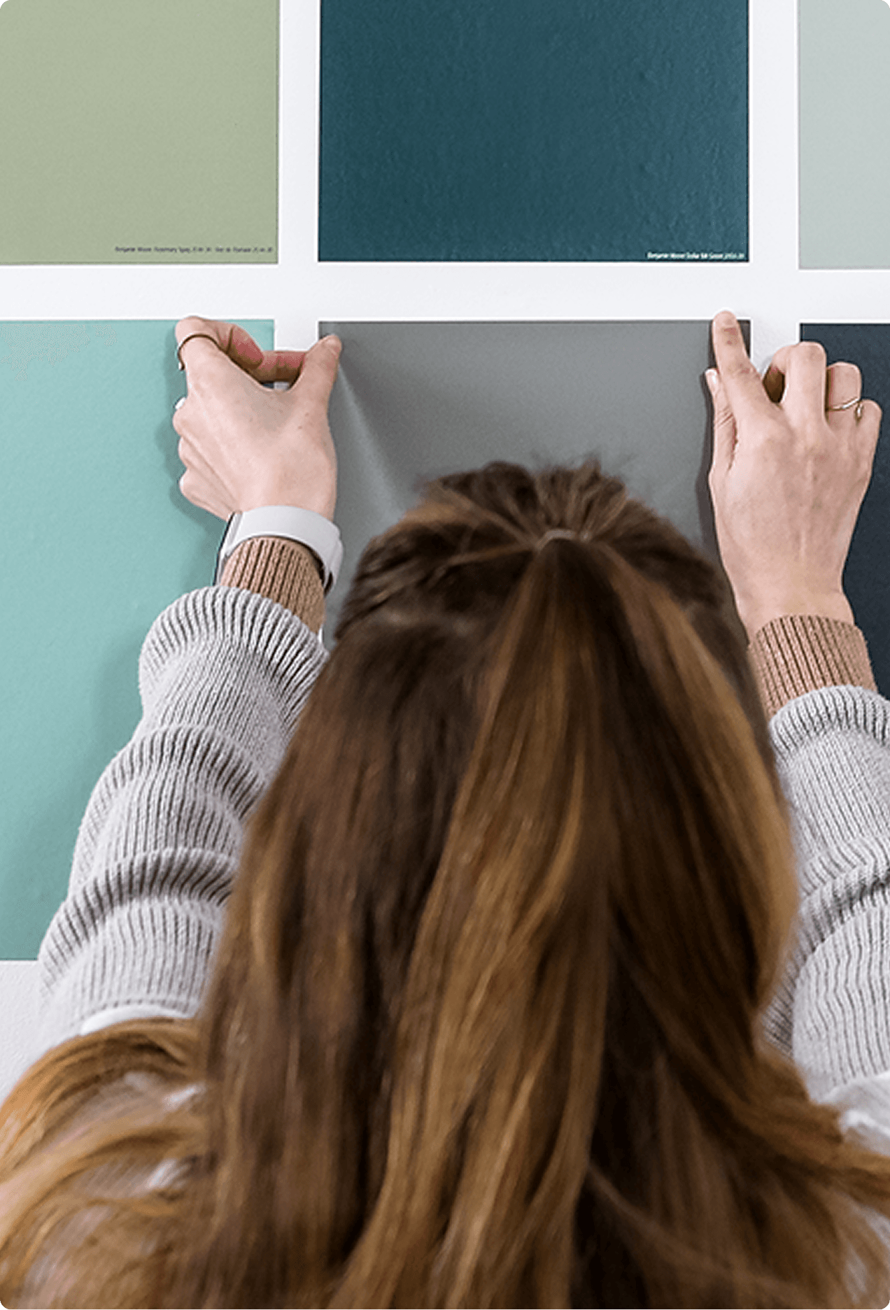 A woman applying Samplize peel-and-stick paint samples to a wall, arranging a grid of green, teal, and neutral color swatches